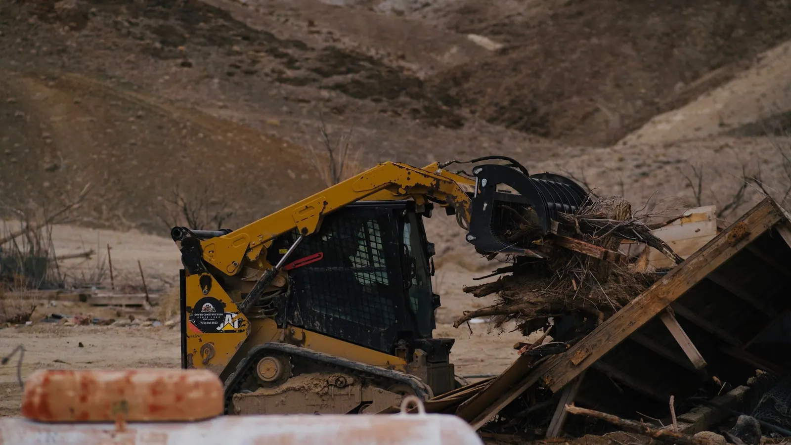 A skid steer grabbing old lumber and debris on a hillside lot