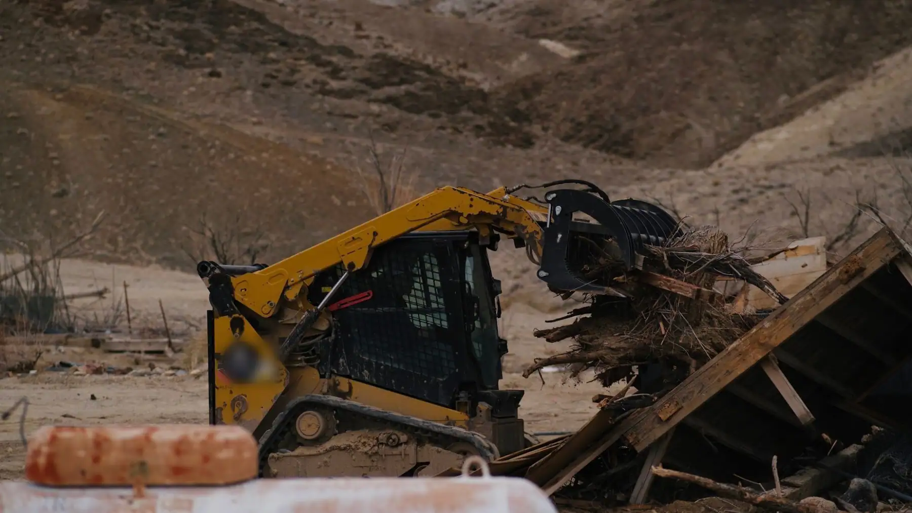 A tracked skid steer with a grapple clearing tree roots and a broken wooden structure on a dirt lot below the hills