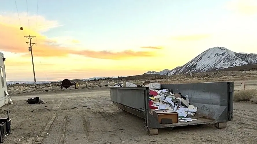 A roll-off dumpster on a dirt lot with a snowy mountain and sunset behind it