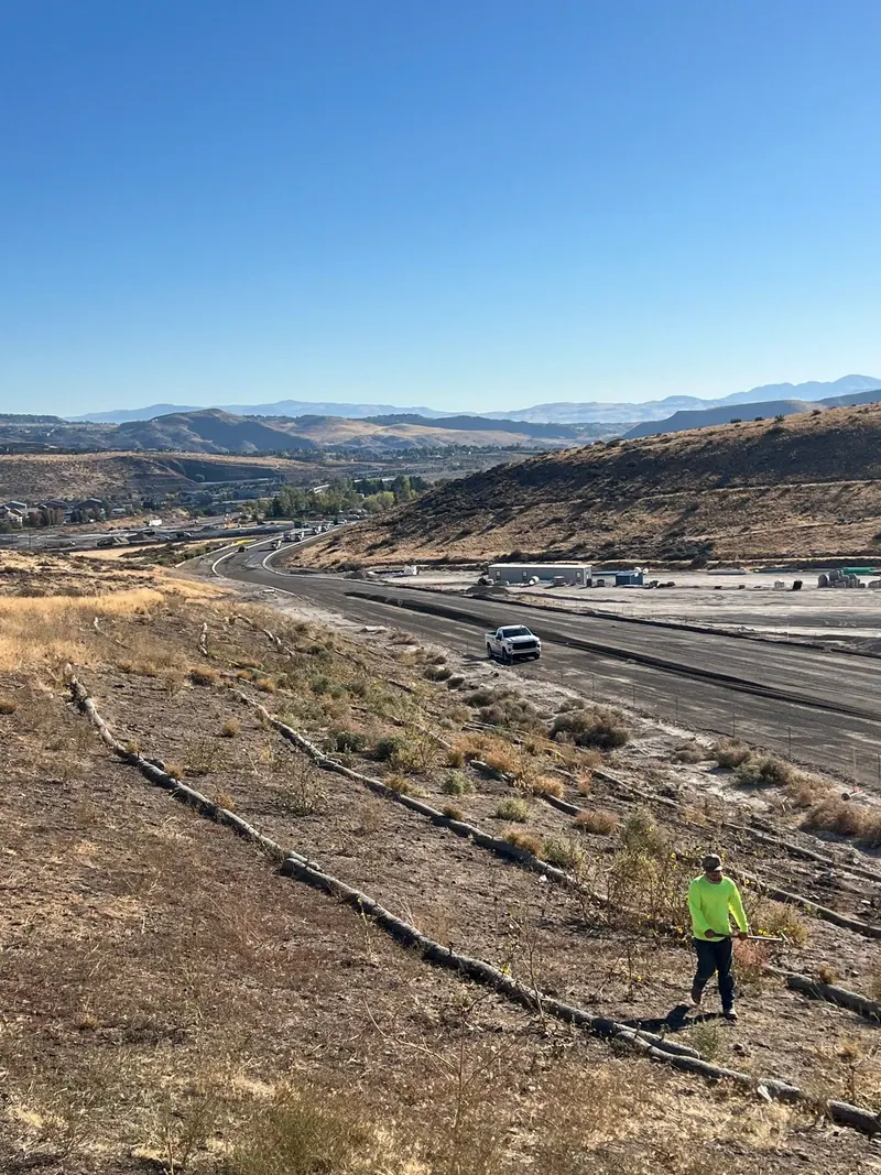 A worker clearing weeds on a long hillside above a highway