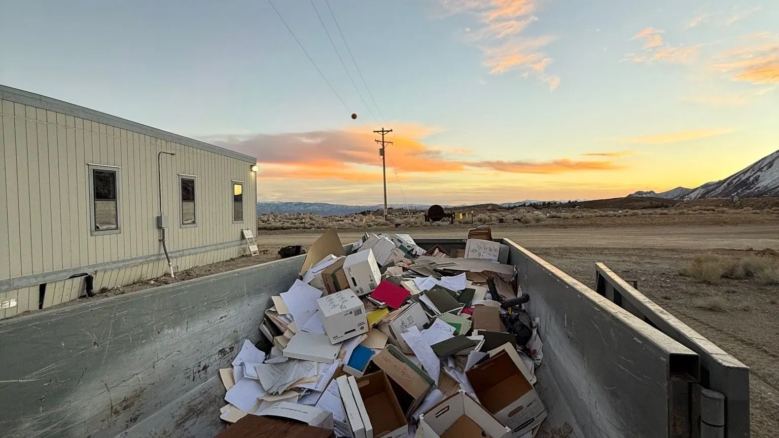 A roll-off dumpster full of boxes beside a commercial building at sunset