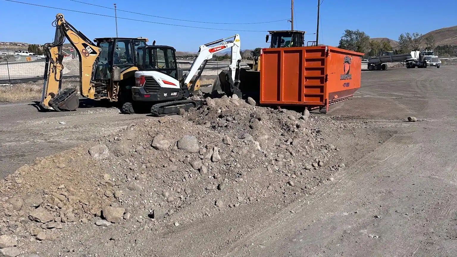 A backhoe and mini excavator beside two roll-off dumpsters on a dirt lot
