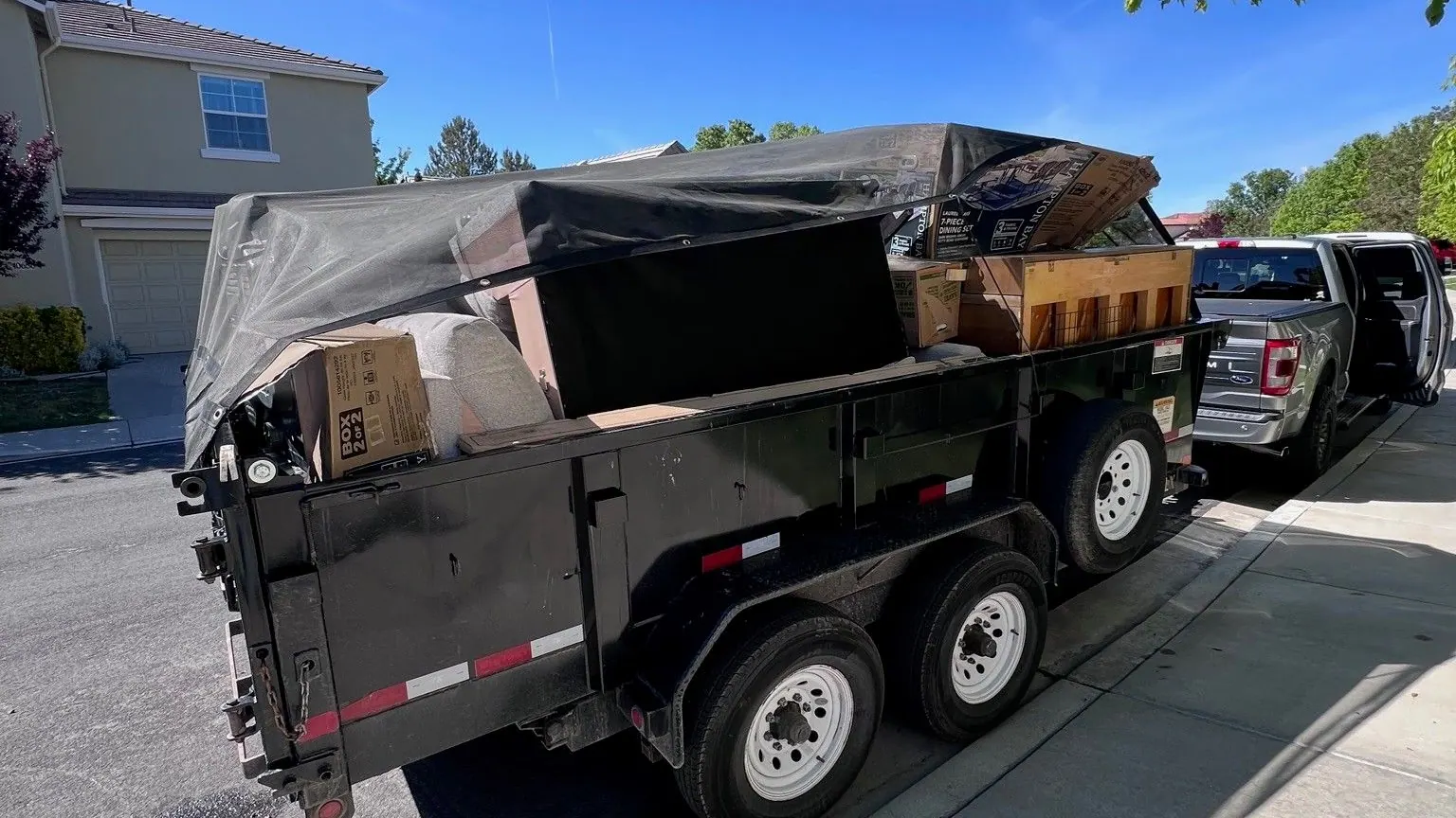 The black dump trailer loaded and tarped at the curb with the pickup ahead of it