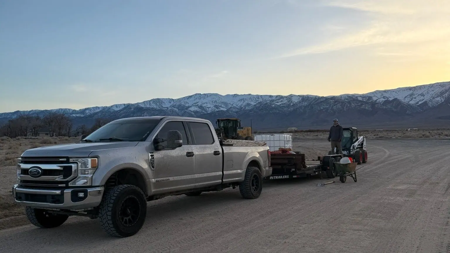 The silver pickup and trailer on an open road at dusk with mountains behind