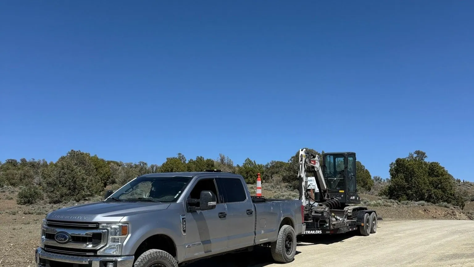 The silver pickup towing a skid steer on a trailer down a dirt road
